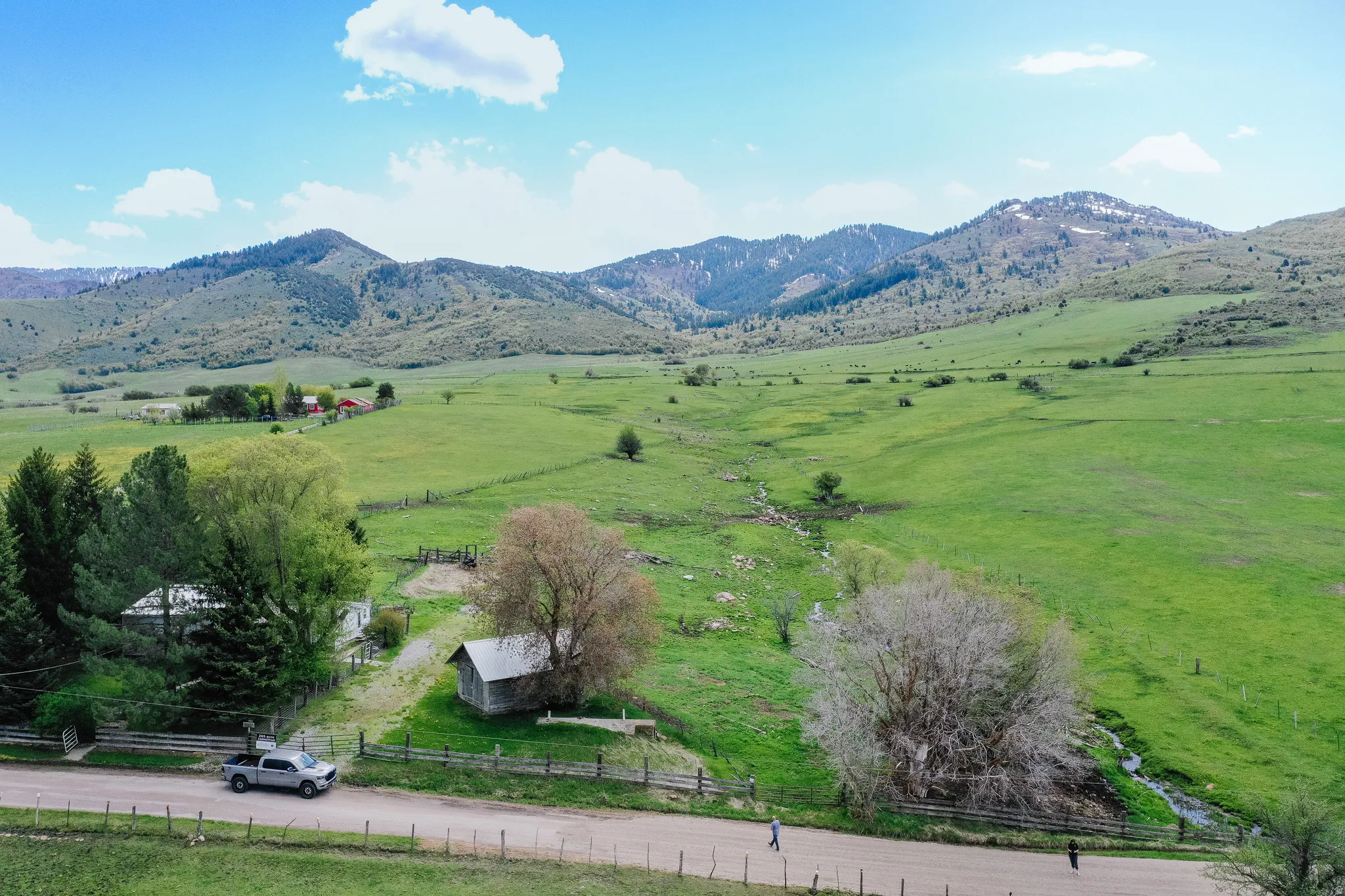 Expansive green landscape with distant mountains, featuring a dirt road, wood fence, and mature trees