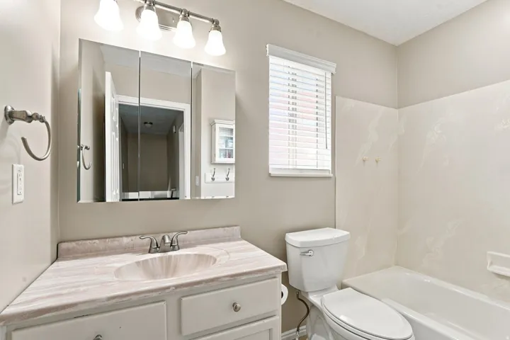Vanity with integrated sink, tri-fold mirrored medicine cabinet, wall-mounted light fixture, white toilet, and tub