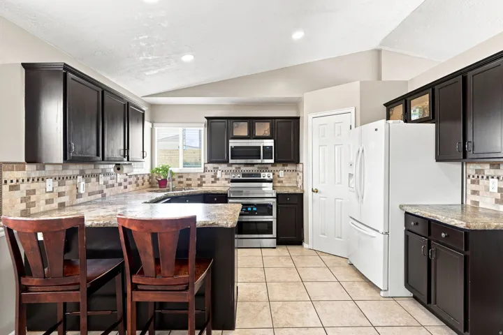 Kitchen featuring dark wood cabinetry, light-colored tile flooring, stone countertops, and a full tile backsplash