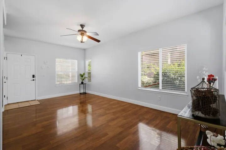 Spacious room featuring gleaming wood-finish flooring, ceiling fan with integrated lighting, multiple windows with plantation shutters providing natural light, white entry door, and neutral wall finishes throughout