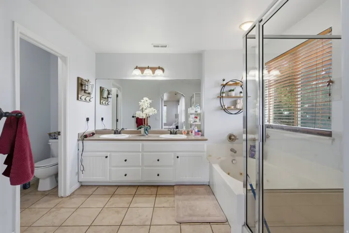 Dual vanity with white cabinetry, tile flooring, built-in soaking tub, glass-enclosed shower, and large wall-mounted mirror