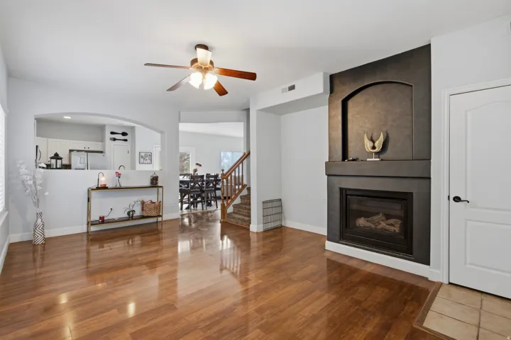 Hardwood-finish flooring, built-in gas fireplace with dark surround, ceiling fan, arched kitchen pass-through, and staircase with wooden handrails