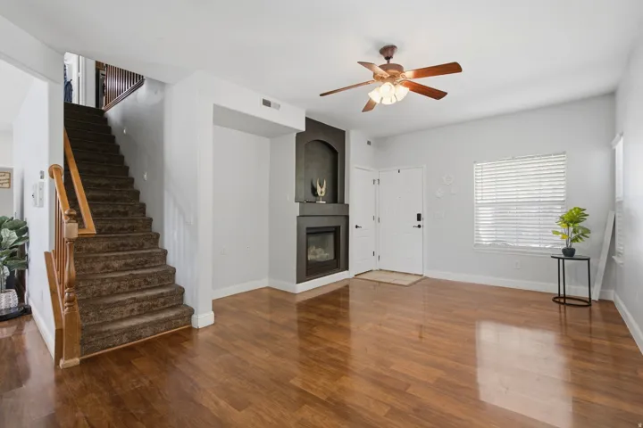 Wood-finish flooring, built-in fireplace, ceiling fan with light fixture, window with blinds, and white entry door