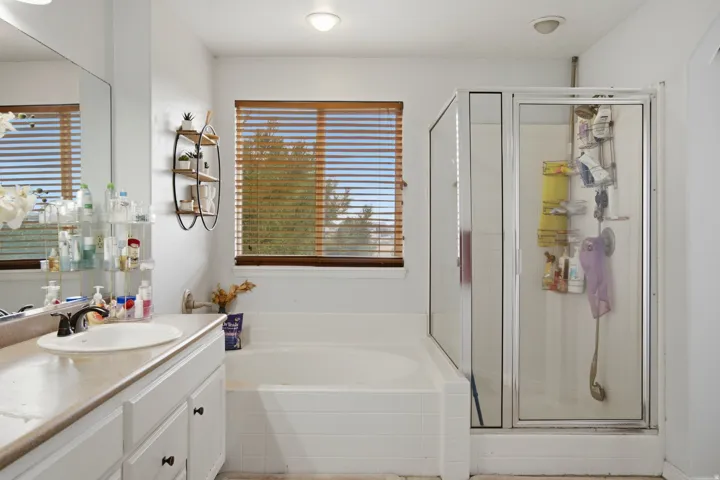Built-in soaking tub with tiled surround, glass-enclosed shower with chrome frame, vanity with an integrated sink and dark hardware, large wall-mounted mirror, and a window with wood-finish blinds