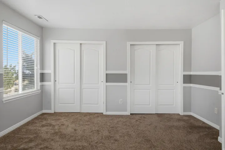 Spacious bedroom with neutral wall finishes, carpeted flooring, abundant natural light from large windows with blinds, and dual closet systems featuring white panel doors with integrated hardware
