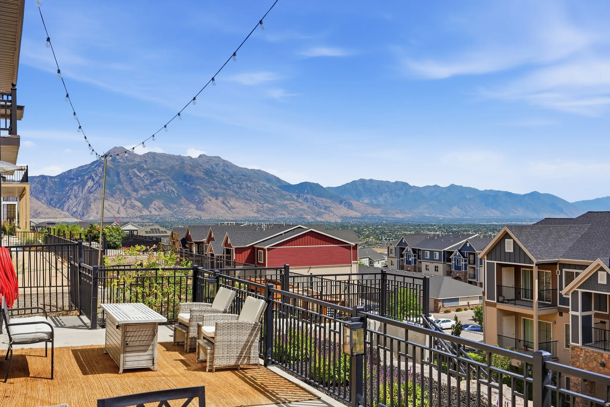 Expansive outdoor patio with a mountain backdrop, featuring a black metal railing, string lighting, and wood-finish flooring