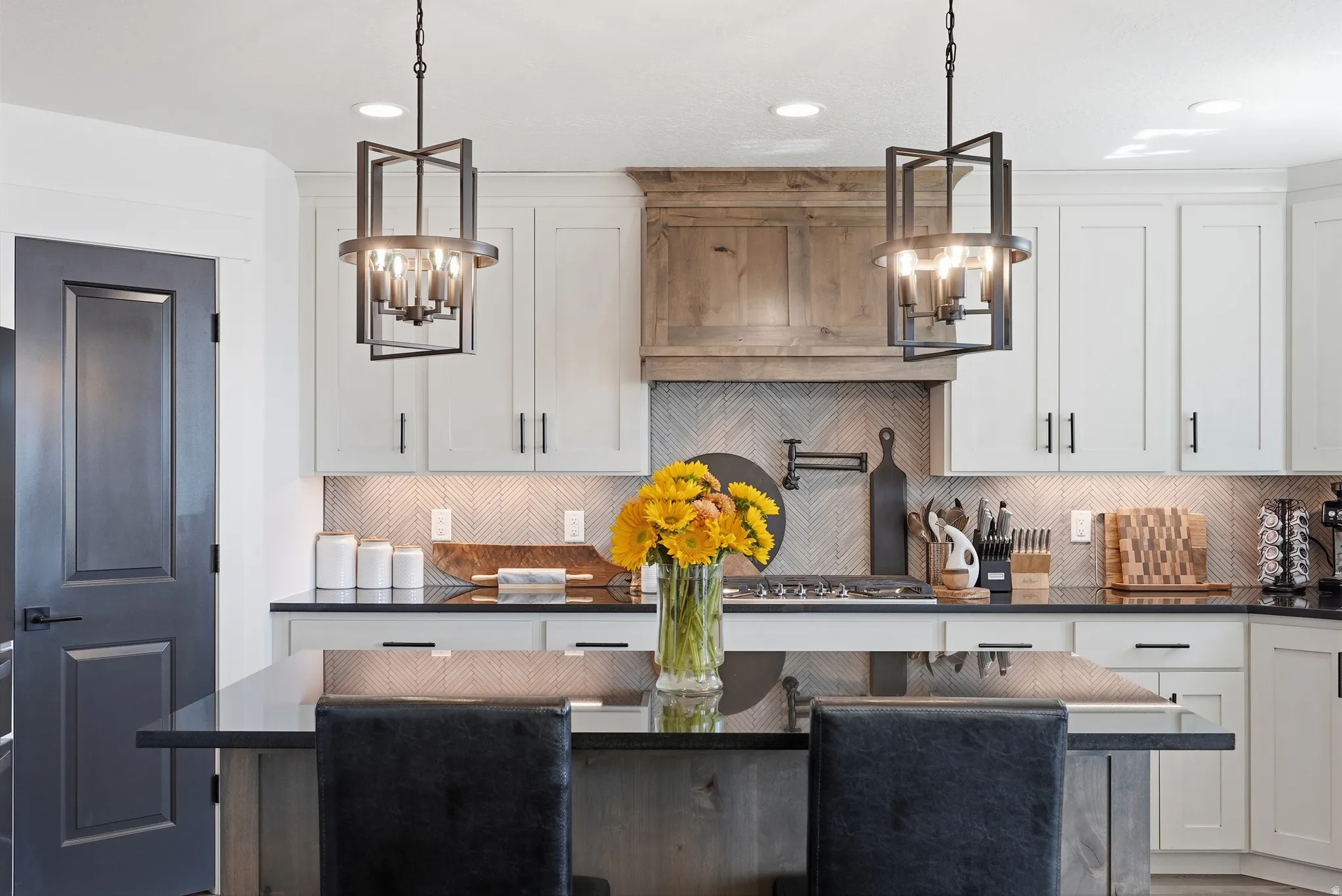 White cabinetry with black hardware, dark countertops, two linear pendant lights, light-toned chevron tile backsplash, and a wooden range hood