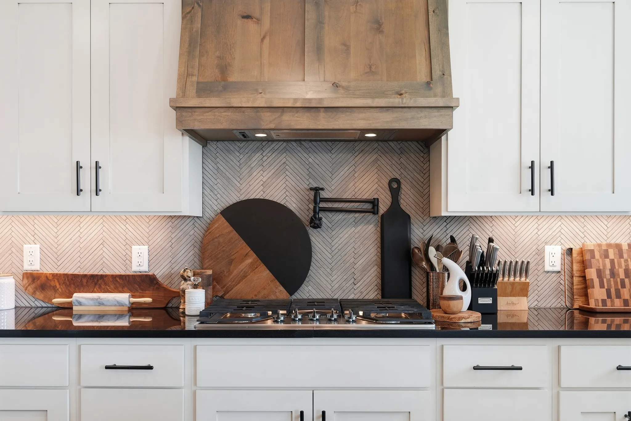 White shaker cabinetry with matte black hardware, wood-finish range hood, gray herringbone tile backsplash, integrated gas cooktop, and dark countertops