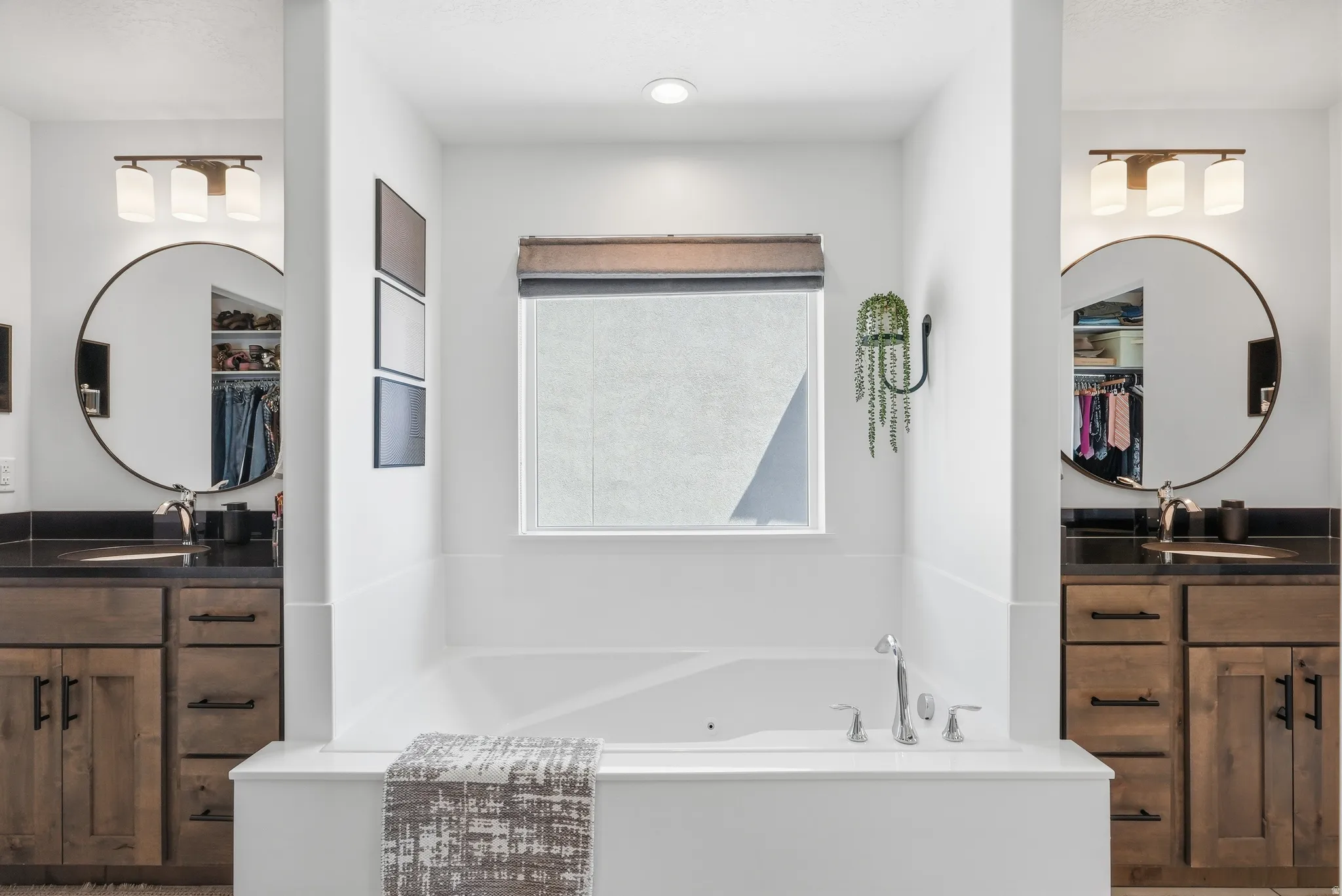 Built-in soaking tub with a window, dual vanity sinks, wood-finish cabinetry, and circular mirrors