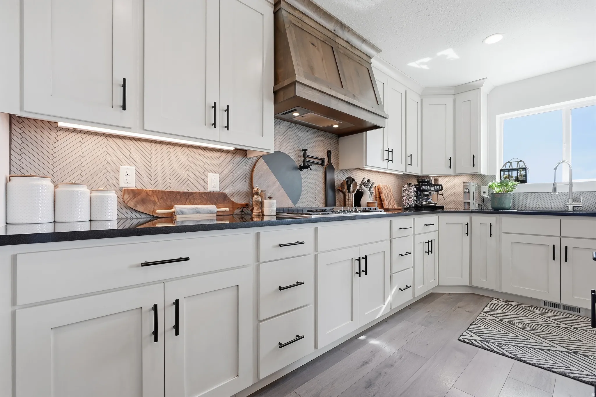 White shaker cabinetry with matte black hardware, dark countertops, a herringbone tile backsplash, wood-finish flooring, and a natural wood range hood