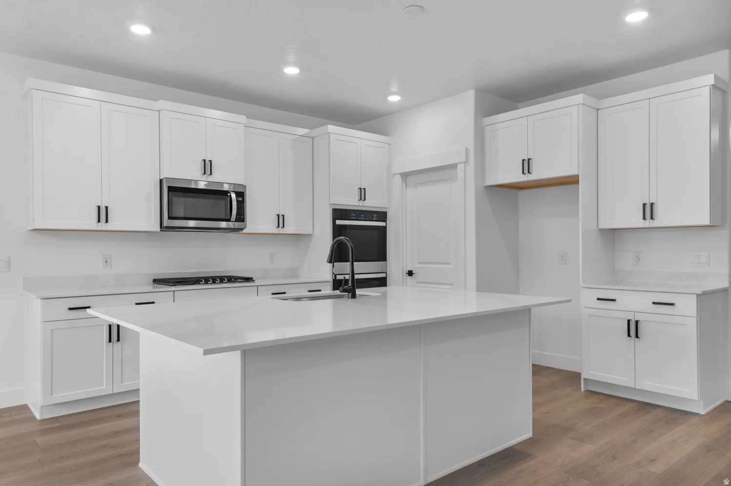 Kitchen featuring white cabinetry, a central island with a sink, stainless steel appliances, light-toned countertops, and wood-finish flooring