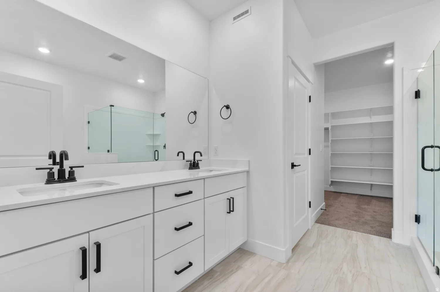 Dual vanity with integrated sinks and black fixtures, expansive wall mirror, glass-enclosed shower, white cabinetry with black hardware, and tiled flooring