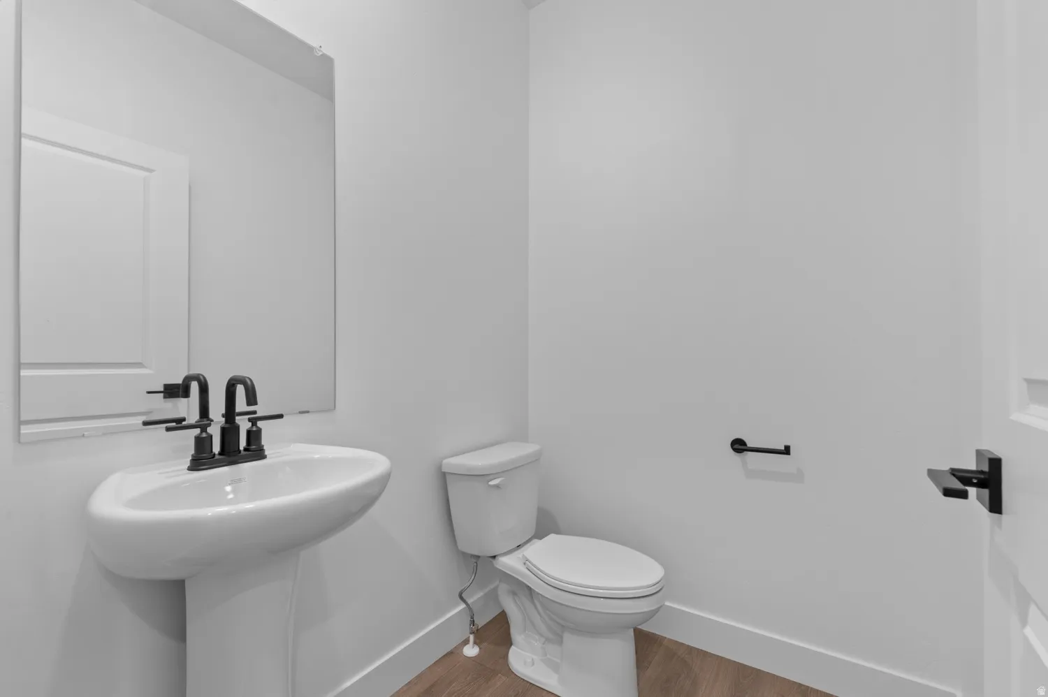 Pedestal sink with matte black faucet, wall-mounted mirror, modern toilet, and wood-finish flooring
