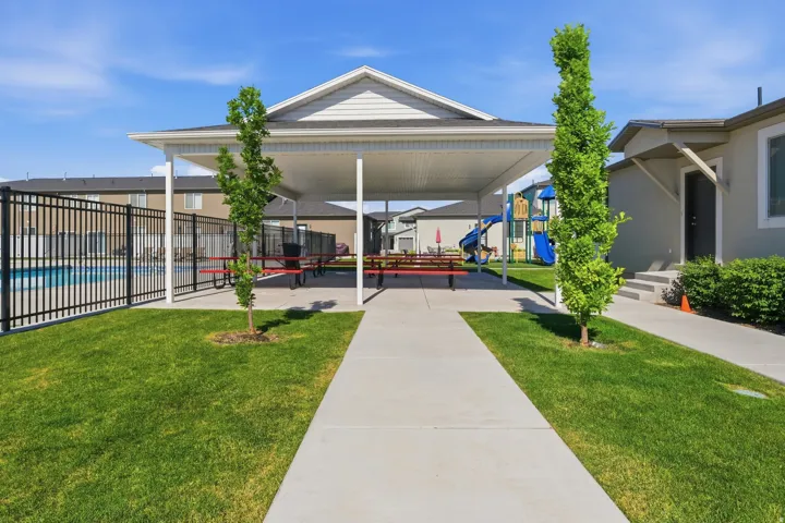 Covered outdoor seating area featuring multiple picnic tables, an adjacent fenced swimming pool, and a nearby playground