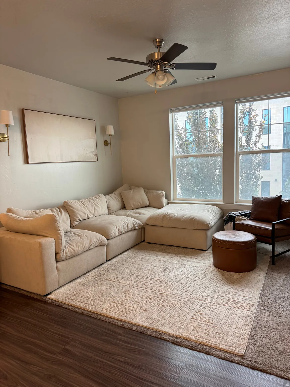 Wood-finish flooring, ceiling fan with integrated lighting, dual wall sconces, and large windows offering natural light
