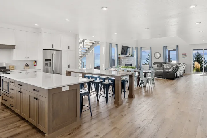 Expansive open-concept living space with light wood-finish flooring, a kitchen island with stone countertops, stainless steel appliances, and white cabinetry