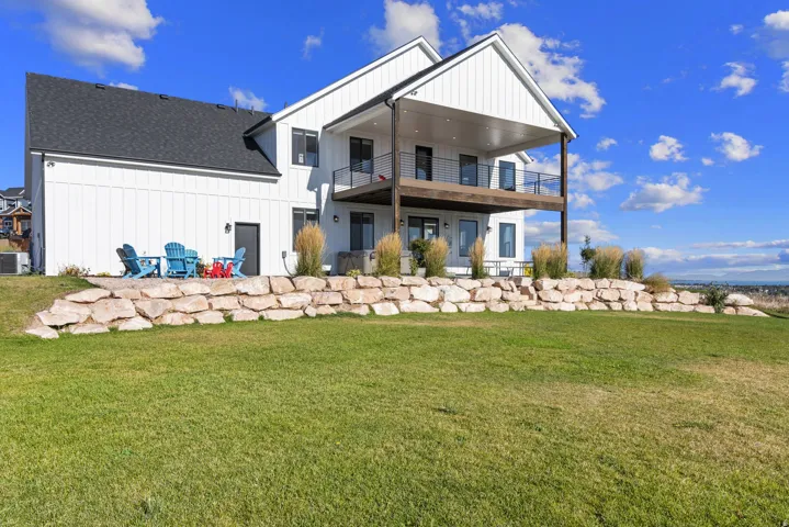 White vertical siding, upper level covered balcony, lower level patio, and a stone retaining wall