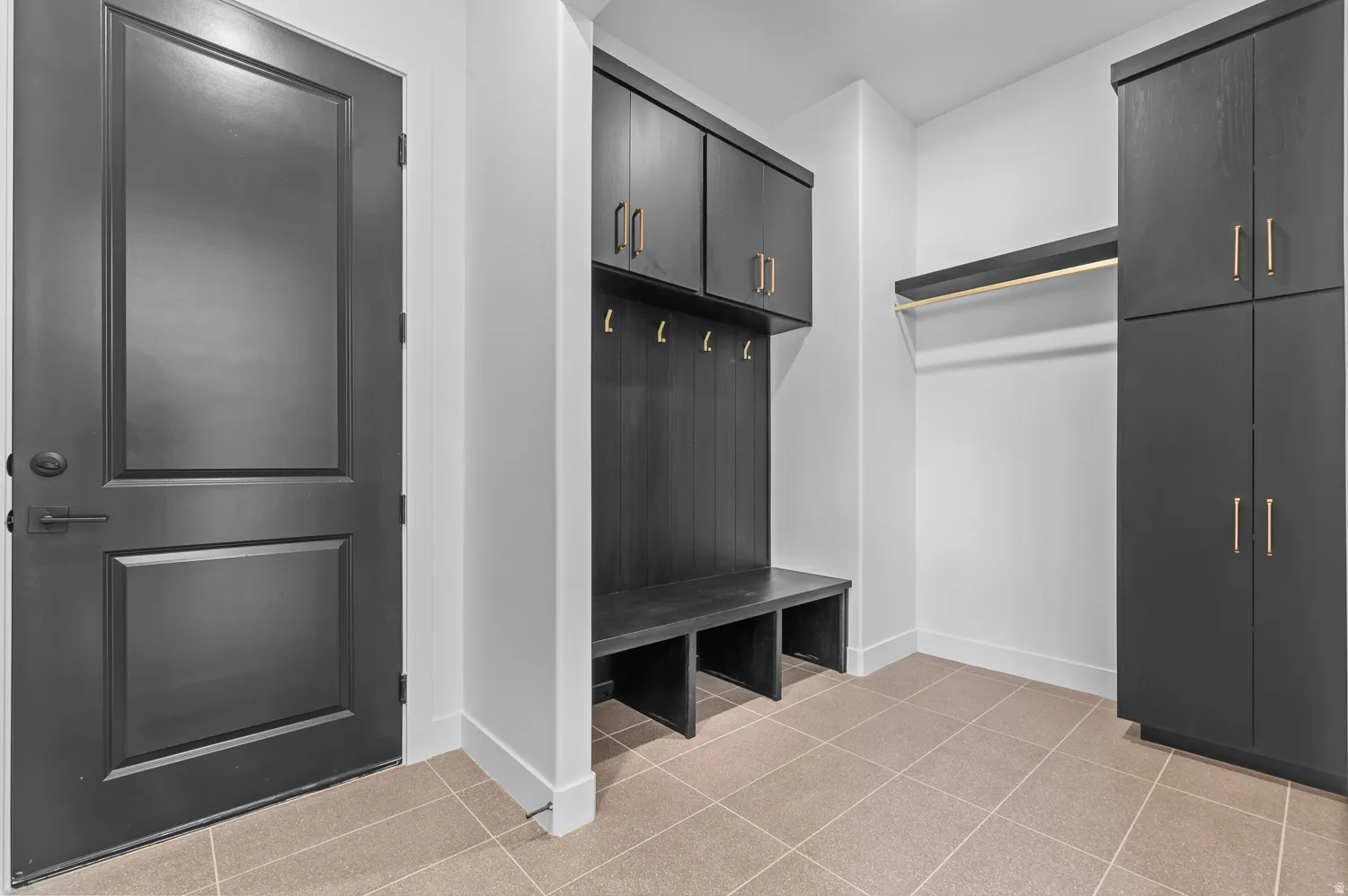 Mudroom featuring light-toned square tile flooring, custom dark wood-finish built-in storage with gold-toned hardware, and a dark paneled door