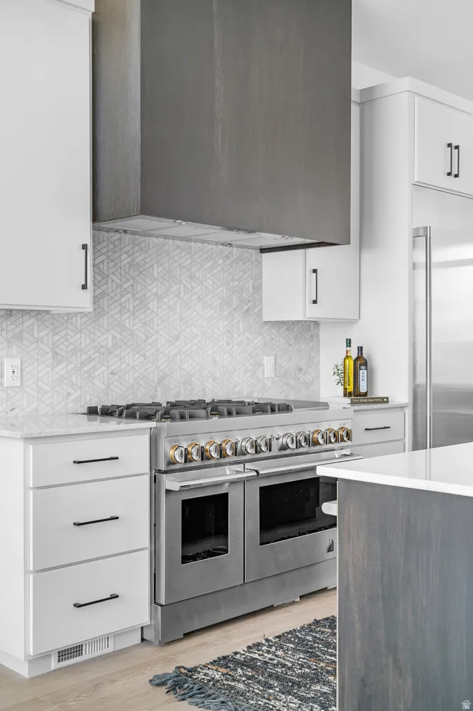 Stainless steel range with gold-toned accents, geometric patterned tile backsplash, custom wood-paneled range hood, white cabinetry with black hardware, and light wood-finish flooring