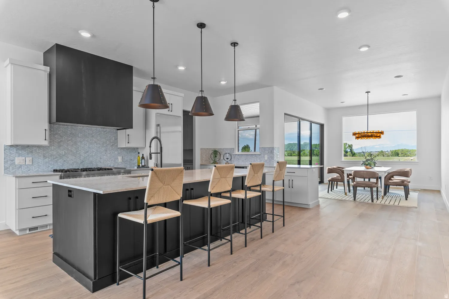 Expansive kitchen island with seating, light wood-finish flooring, custom range hood, patterned tile backsplash, and large windows with outdoor scenery