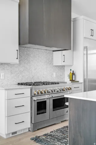 Stainless steel range with gold-toned accents, geometric patterned tile backsplash, custom wood-paneled range hood, white cabinetry with black hardware, and light wood-finish flooring