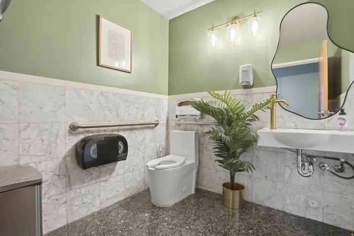 White marble-look wall tiling, modern wall-mounted sink with gold-tone faucet, gold-tone vanity light fixture, and contemporary frameless mirror