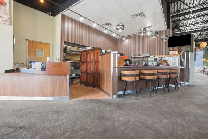 Textured gray carpet flooring, wood-finish reception desk, wood-finish bar with seating, industrial exposed ceiling, and recessed and pendant lighting fixtures