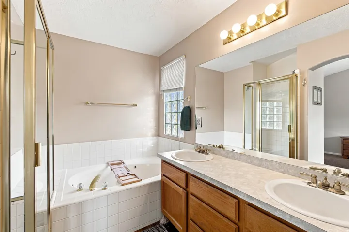 Dual vanity with integrated sinks, a built-in jetted tub, a separate shower enclosure with a glass door, and a large mirror with overhead lighting