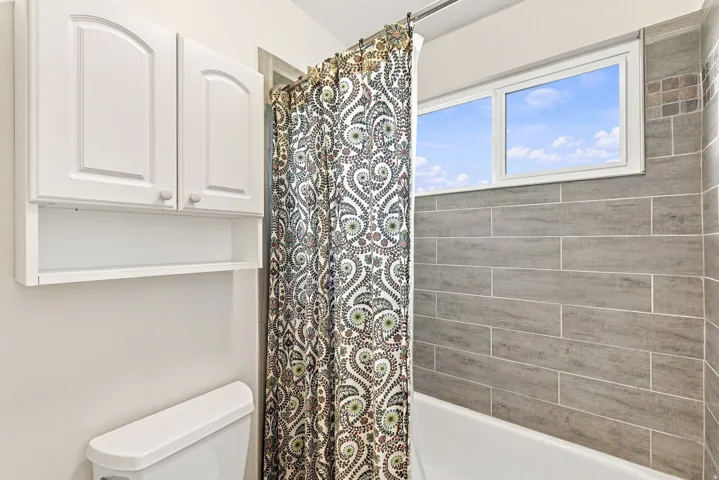 White wall-mounted cabinetry, white toilet, white framed window, and gray tiled shower surround with accent tiles