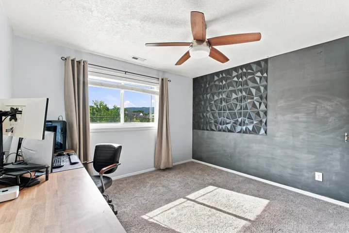 Carpeted flooring, multi-pane window, ceiling fan with light fixture, and a textured accent wall