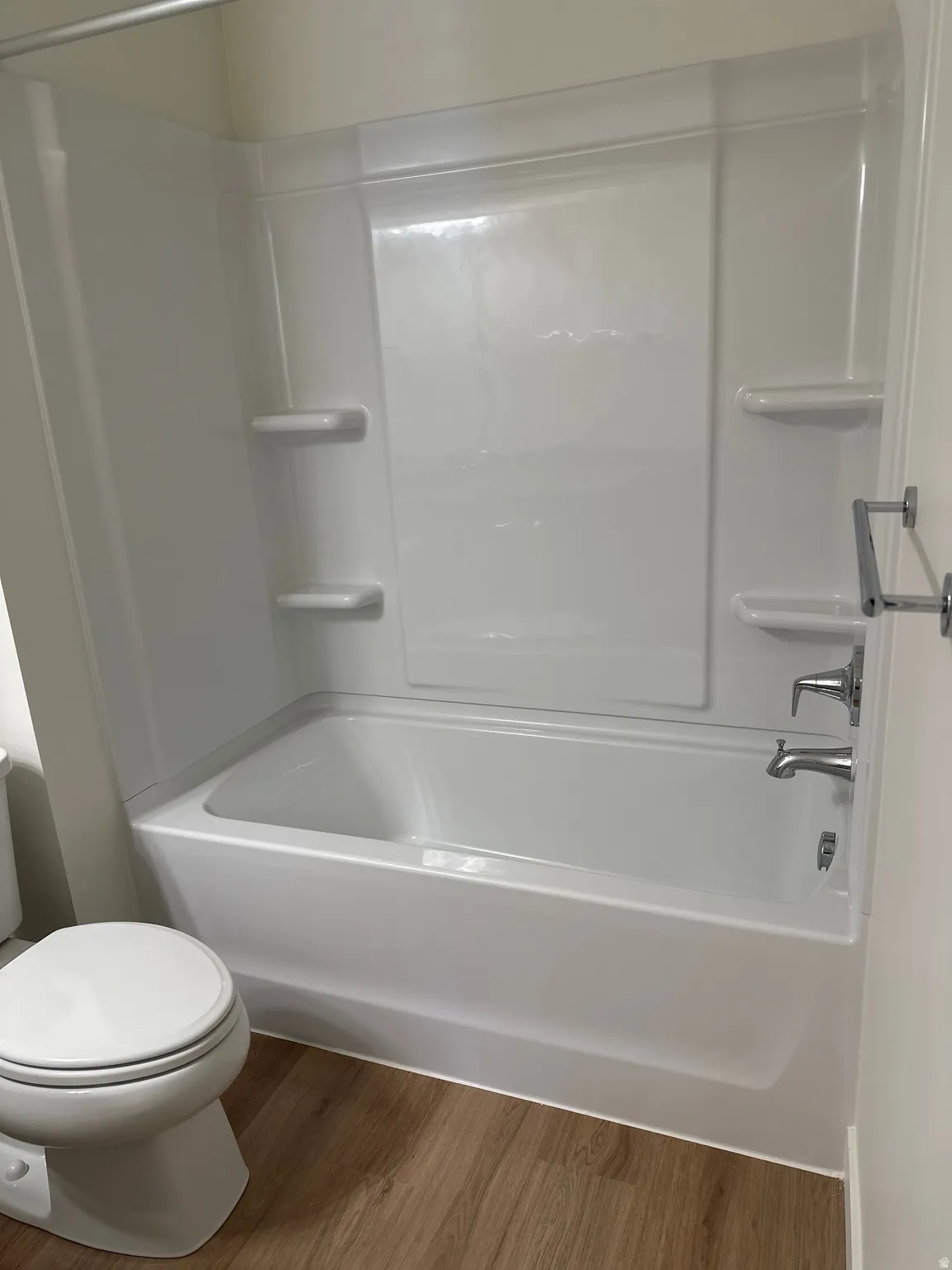 White bathtub with integrated shower surround, chrome fixtures, and wood-finish flooring