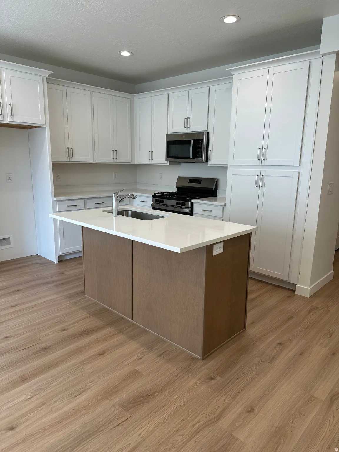 White shaker-style cabinetry, stainless steel appliances, light wood-finish flooring, white countertops, and a kitchen island with an integrated sink