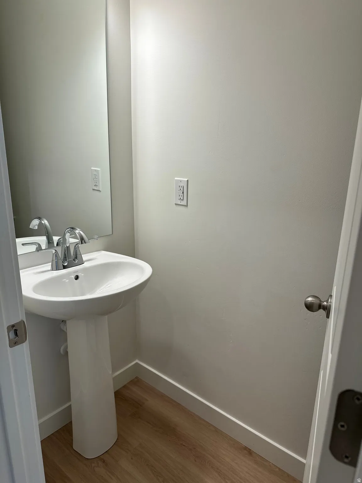 Pedestal sink with a chrome faucet, wall-mounted mirror, and wood-finish flooring