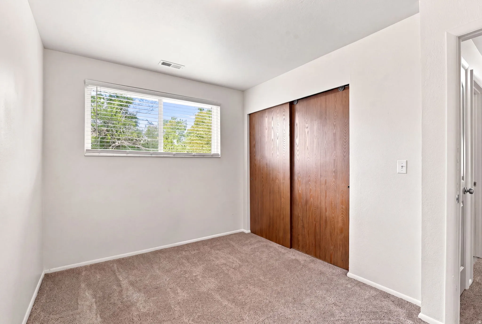 Wall-to-wall carpeting with a neutral finish, a single window, and natural wood-finish sliding closet doors