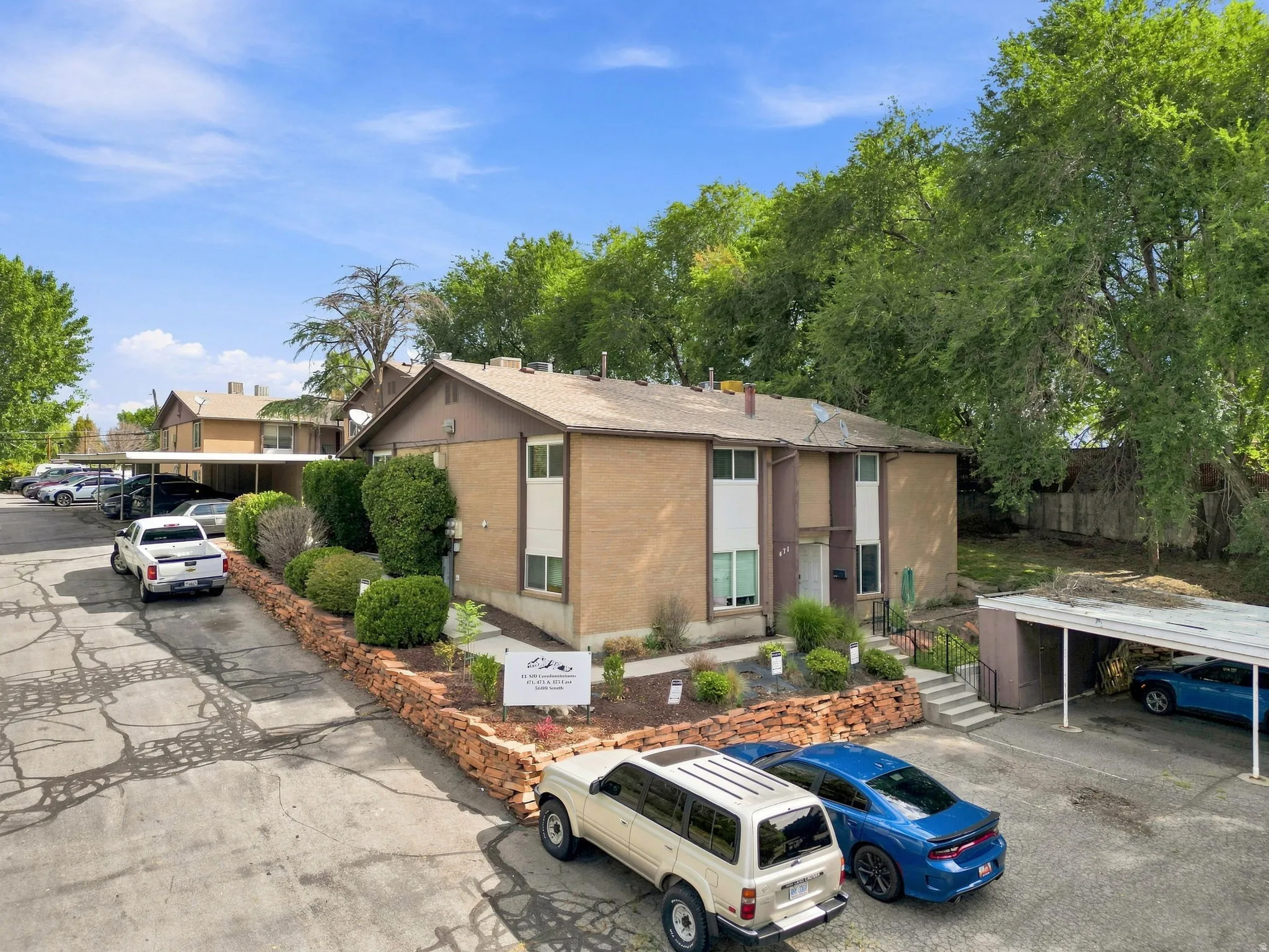 Brick and stucco exterior with multiple units, covered parking, mature landscaping, and an organized parking area