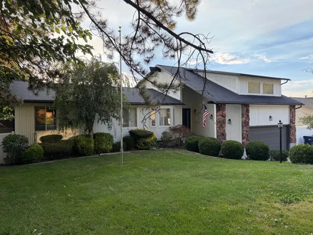 Exterior featuring a pitched roof, stone pillar accents, white vertical siding, an attached garage, and a landscaped front yard with a manicured lawn