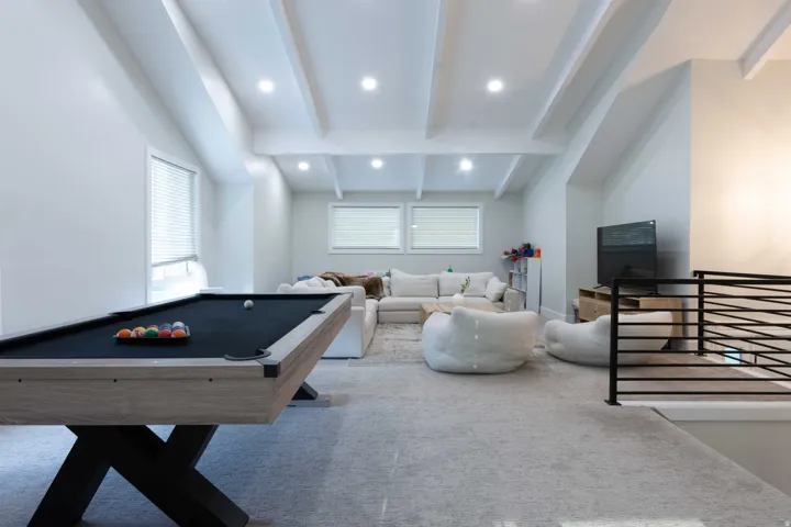 Vaulted ceiling with exposed beams, recessed lighting, multiple windows with blinds, and light-toned carpeting