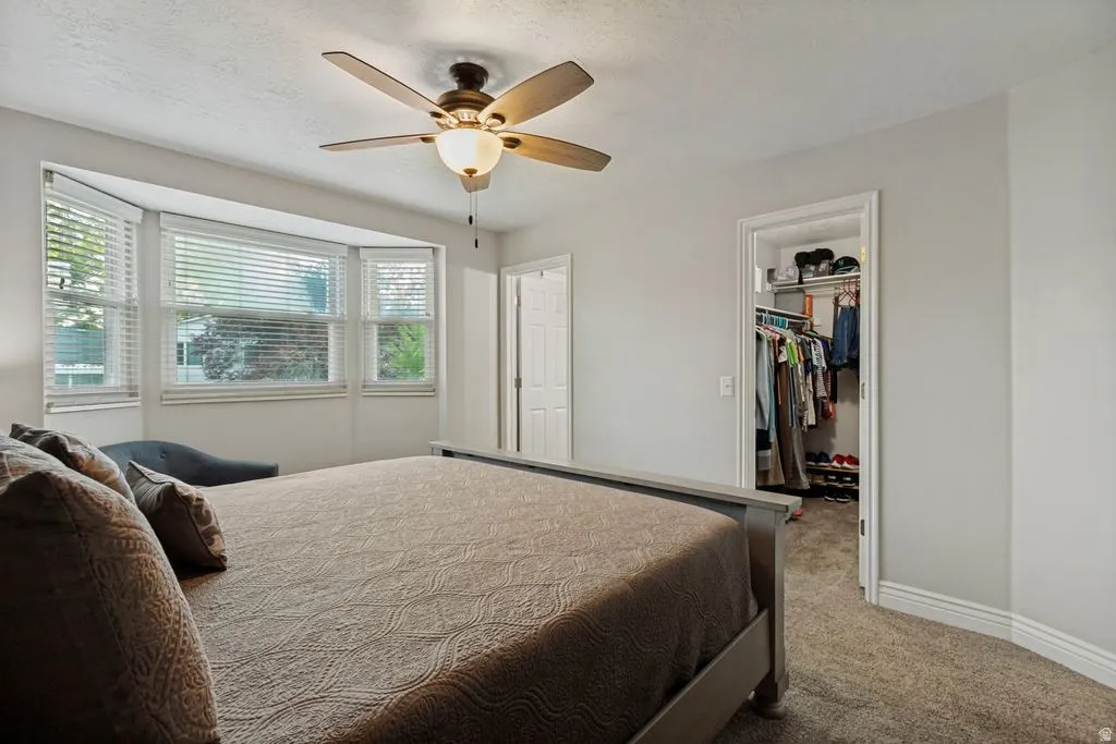 Bay window with horizontal blinds, ceiling fan with integrated lighting, recessed closet with built-in shelving, white trim, and carpeted flooring