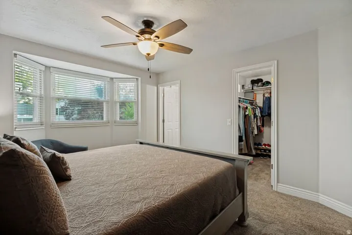 Bay window with horizontal blinds, ceiling fan with integrated lighting, recessed closet with built-in shelving, white trim, and carpeted flooring