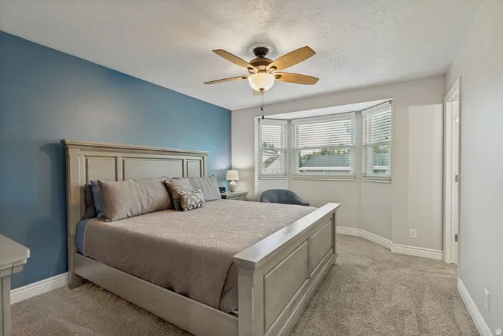 Neutral carpet flooring, a ceiling fan with integrated lighting, and a bay window with blinds