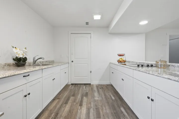 White cabinetry with black hardware, wood-finish flooring, light-toned countertops, integrated cooktop, and a stainless steel sink with a high-arc faucet