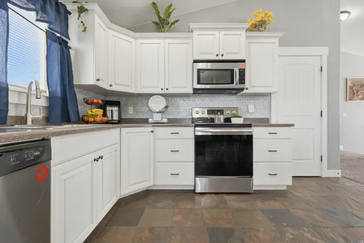 White cabinetry, stainless steel appliances, patterned tile backsplash, dark countertops, and slate-finish flooring