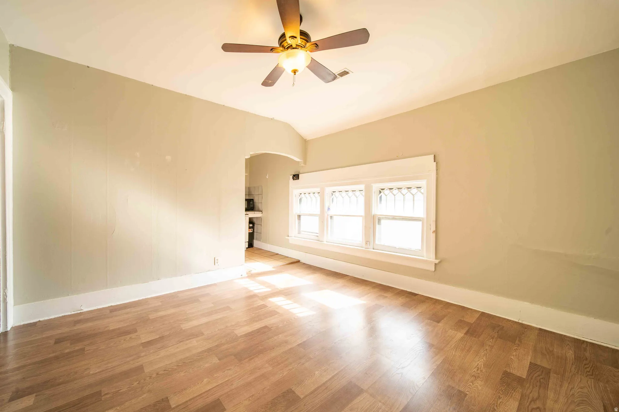 Wood-finish flooring, ceiling fan with integrated lighting, multiple windows with decorative grilles, and an arched doorway