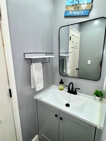 Integrated vanity with white countertop and sink, gray cabinetry, matte black faucet, and a wall-mounted mirror with a rounded black frame