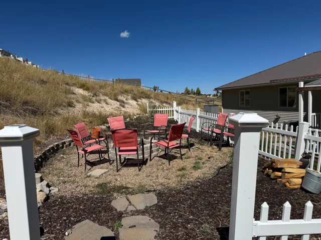 Outdoor fire pit area with stone edging and surrounding ground cover, enclosed by a white picket fence, featuring a hillside backdrop