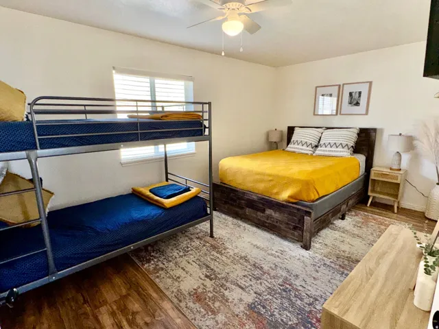 Wood-finish flooring, ceiling fan with integrated lighting, window with horizontal blinds, and a built-in wall-mounted television
