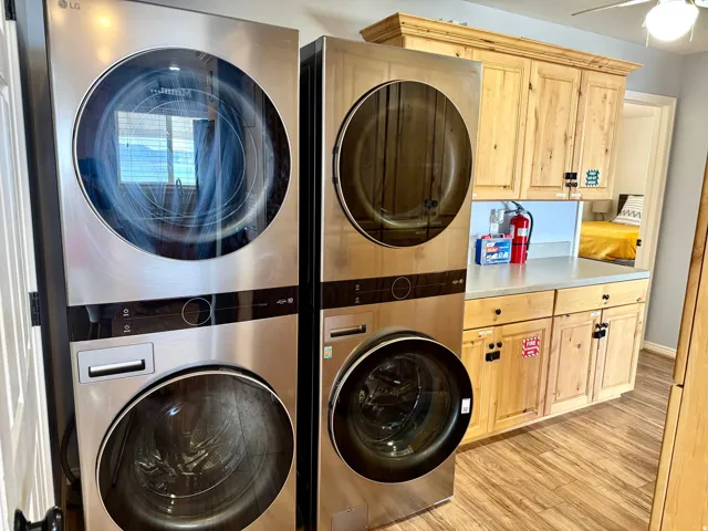 Stacked stainless steel laundry appliances, wood-finish flooring, and natural wood cabinetry with a light-colored countertop