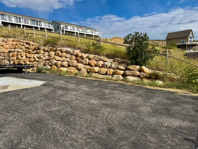 Paved surface with a natural stone retaining wall, mature trees, and distant residential structures