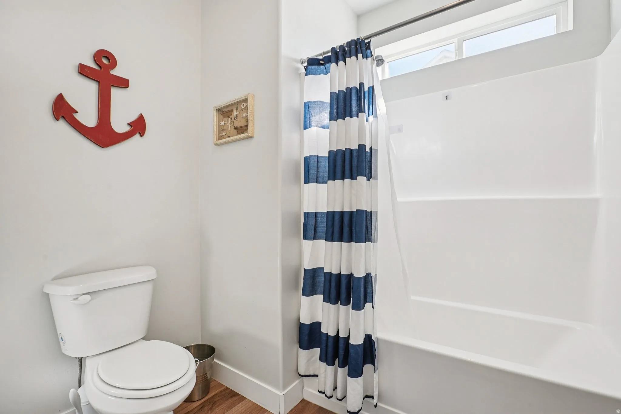 White toilet with a built-in tank, a white tub and shower combination, and wood-finish flooring
