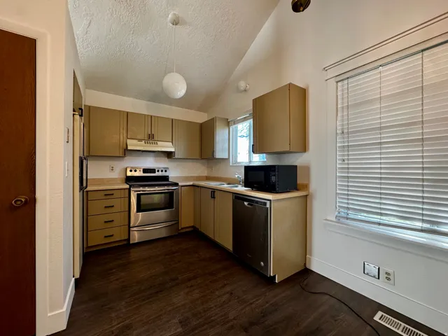 Sloped ceiling kitchen featuring wood-finish flooring, light-toned cabinetry, stainless steel appliances, and a window with blinds